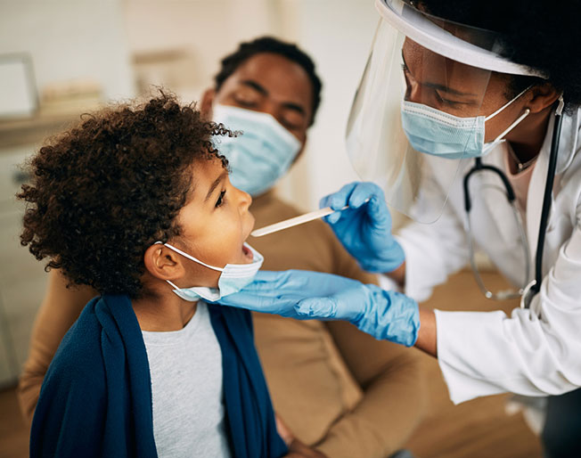 masked doctor places tongue depressor in child's mouth