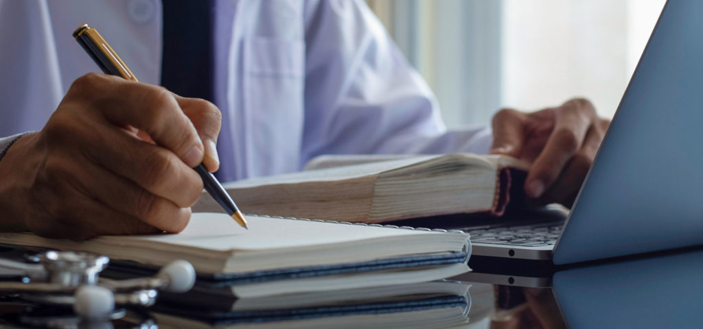 doctor takes notes on legal pad while consulting book and computer