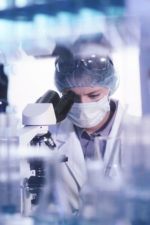 masked lab technician looks into microscope