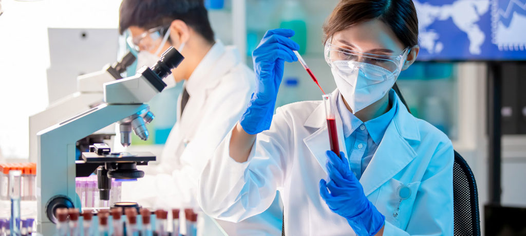 one lab scientist looks through a microscope, another places liquid in vial from syringe