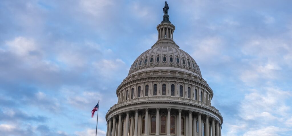 The dome of the United States Capitol building with an American flag waving beside it, set against a partly cloudy blue sky